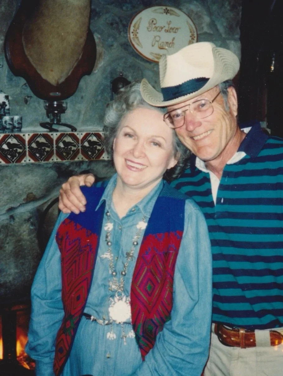 Bob and Fae Hardy smiling beside the stone fireplace at the ranch, the Poco Loco Ranch plaque set in the rock above them